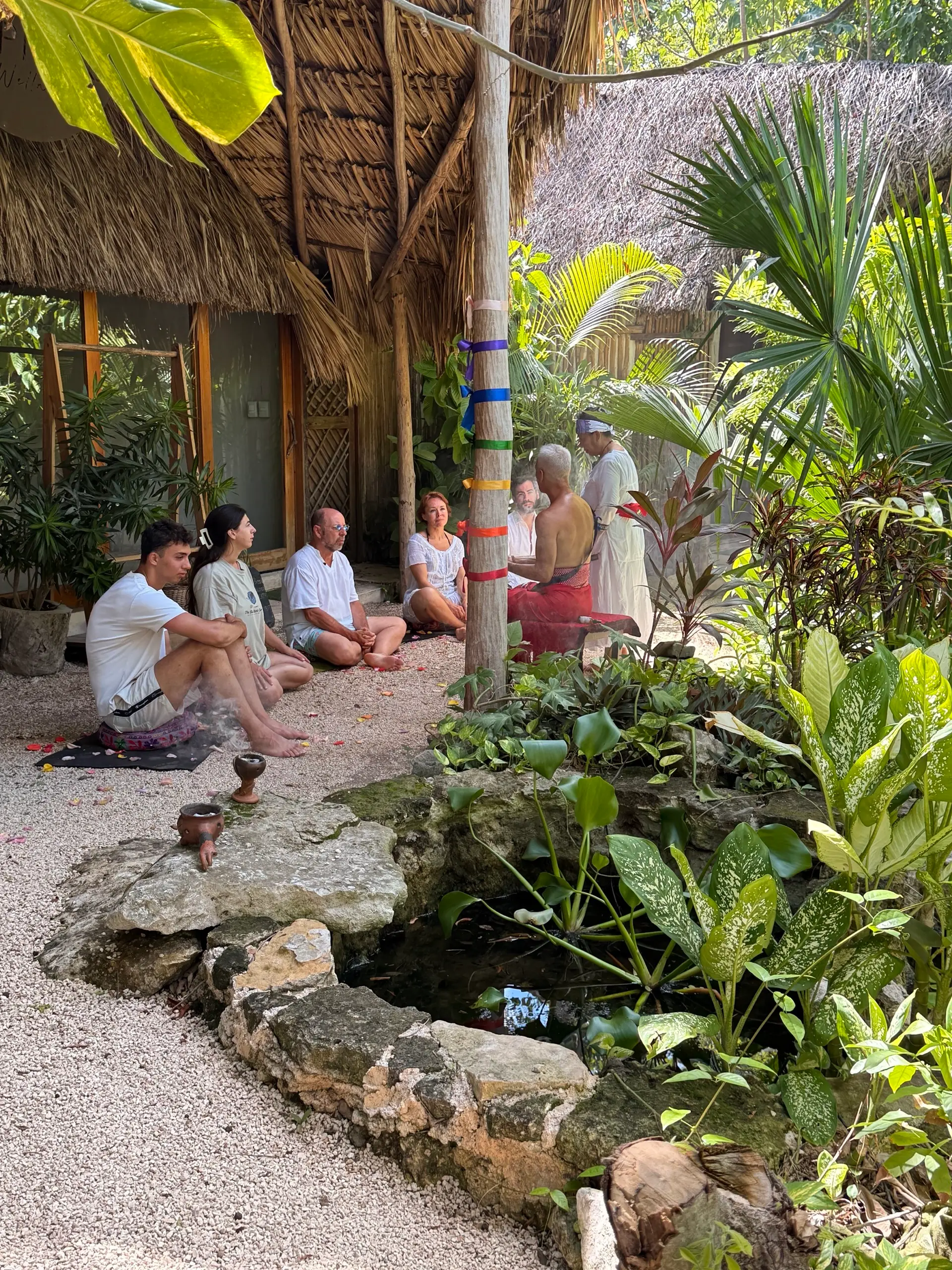 A circle of people sitting under the palapa, beside the ribboned post